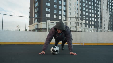 businessman performing push-ups alternating hand placement on soccer ball demonstrating strength, in outdoor urban arena with goal post and residential buildings in the background