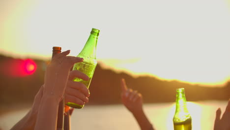 los estudiantes jóvenes están bailando con las manos en alto y haciendo sonar la cerveza de las botellas de colores. esta es una fiesta loca al atardecer en la costa del río.