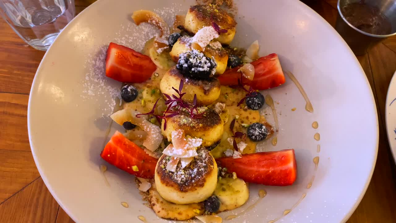 delicioso syrniki con bayas del bosque, virutas de plátano y coco en crema de anacardo y miel, deliciosos mini panqueques de queso para el desayuno, plato tradicional ruso ucraniano, tiro de 4k