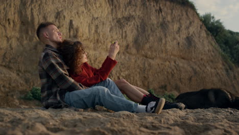 Happy-man-and-woman-relaxing-on-beach.-Cheerful-girl-taking-photo-on-smartphone