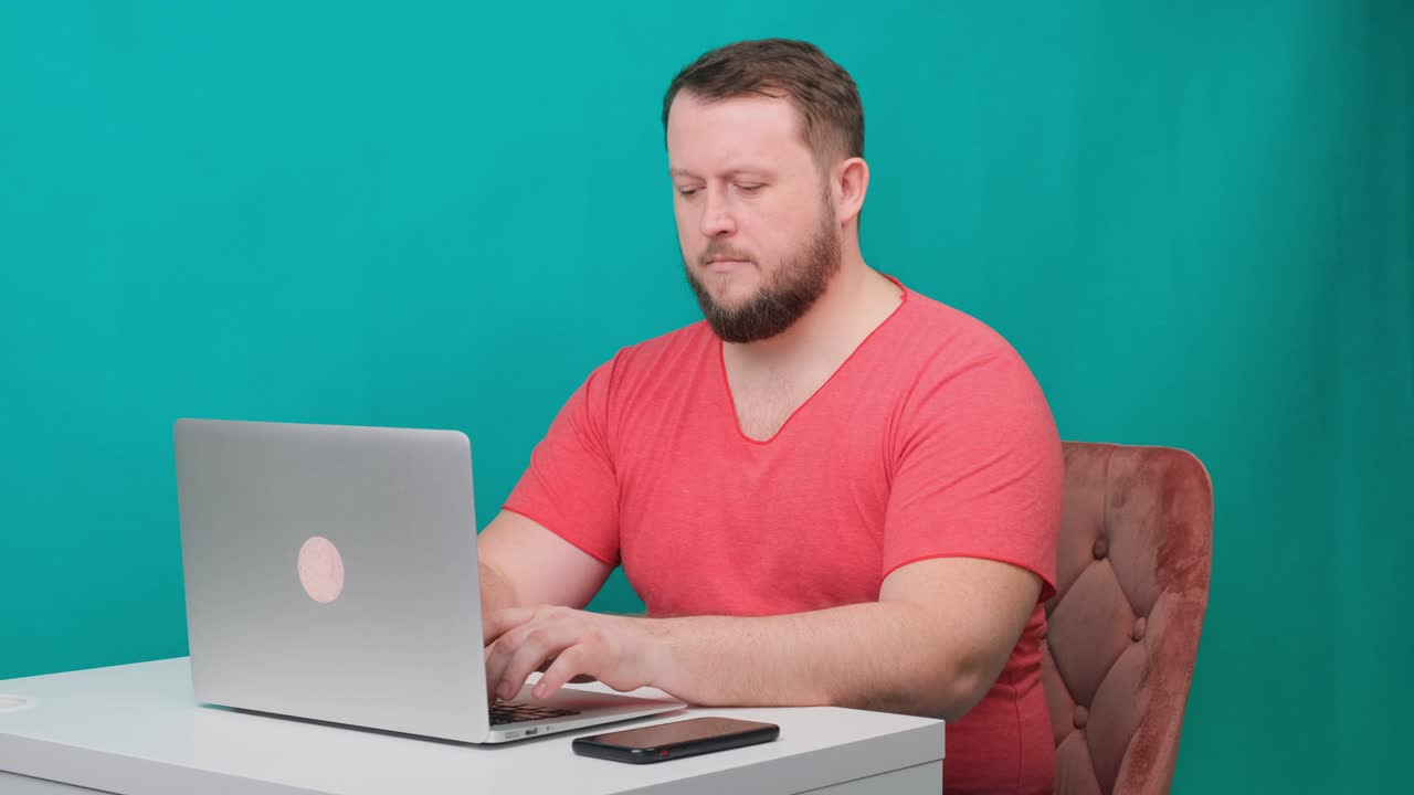 분홍색 티셔츠를 입은 행복한 젊은 사업가가 녹색 화면의 노트북에서 일합니다. 노트북을 바라보는 남성 이야기하는 남자의 초상화. 사무실에서 그의 책상에서 일하는 남자.