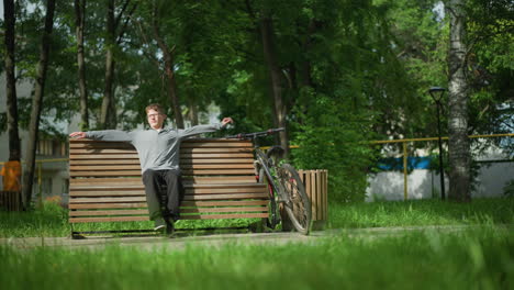 young boy stretches on park bench, then glances back at his bicycle, nearby, someone sits on another bench, and others stroll through the tree-lined park, with greenery in the background