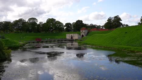 Gente-Cruzando-Un-Puente-Hacia-La-Puerta-De-La-Fortaleza-De-Kastellet-Junto-A-Un-Estanque-Con-Reflejo-Del-Cielo,-Copenhague