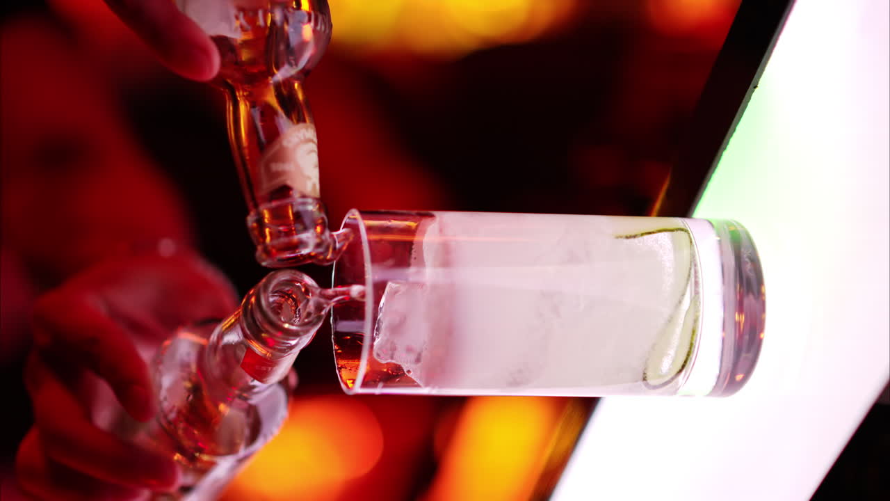 Barman pouring soda into an ice glass at the bar at night with red neon lights, slow motion vertical screen