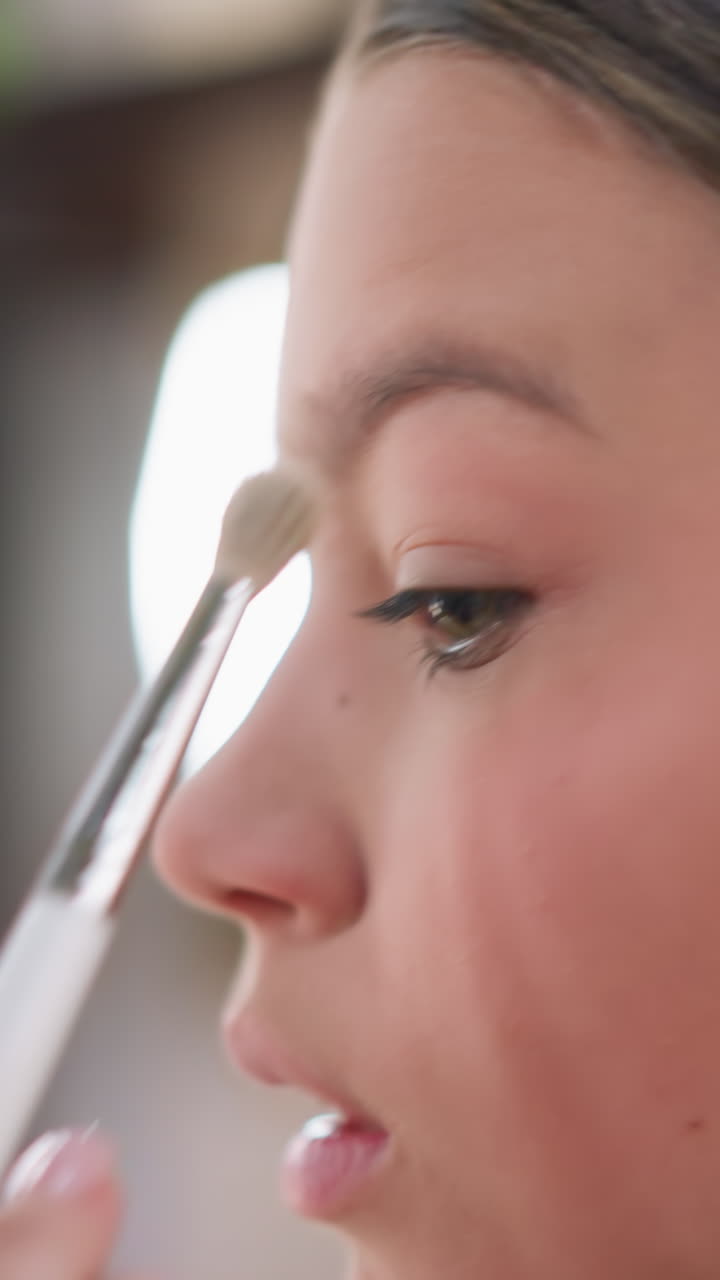 Primer plano de una chica joven aplicándose sombra de ojos con un pincel con una iluminación natural suave, perfil, trazos delicados del pincel a lo largo del párpado y la línea inferior de las pestañas, expresión tranquila y concentrada, fondo con efecto bokeh y tocador, tonos melocotón suaves
