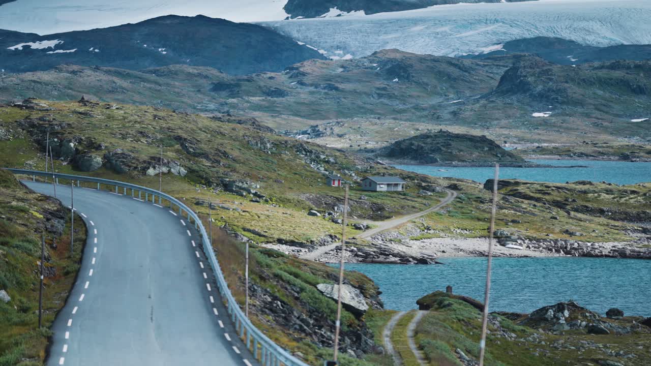 A scenic mountain road winds through a rocky landscape dotted with lakes, cabins scattered throughout the valley, and a glacier dominating the background. Parallax shot.