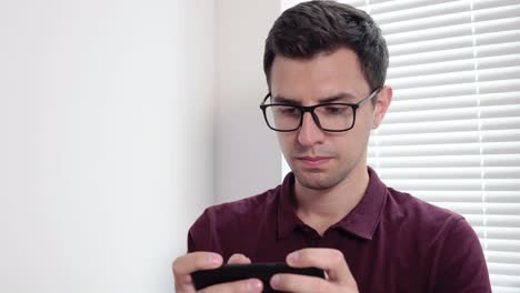 Man-sms-texting-using-app-on-smart-phone-in-the-office-standing-by-the-window.-Handsome-young-business-man-using-smartphone-wearing-glasses-indoors.-Shot-in-4k