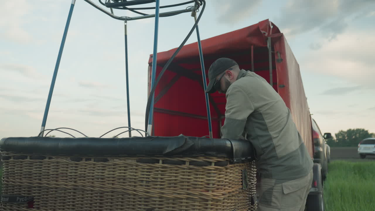close up of woman bending to secure gas cylinder handed by man into wicker balloon basket on grassy field under cloudy sky, focus on careful transfer and safety prep before hot air flight