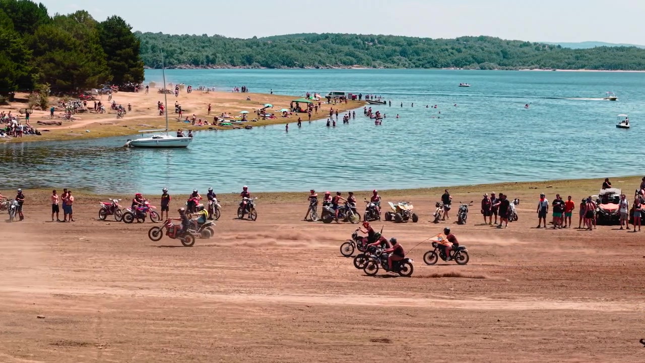 Motorcyklister kører på crossere på en støvet strand nær en sø