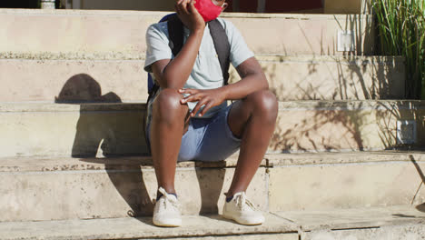 sad african american boy wearing face mask sitting on stairs outdoors on a sunny day