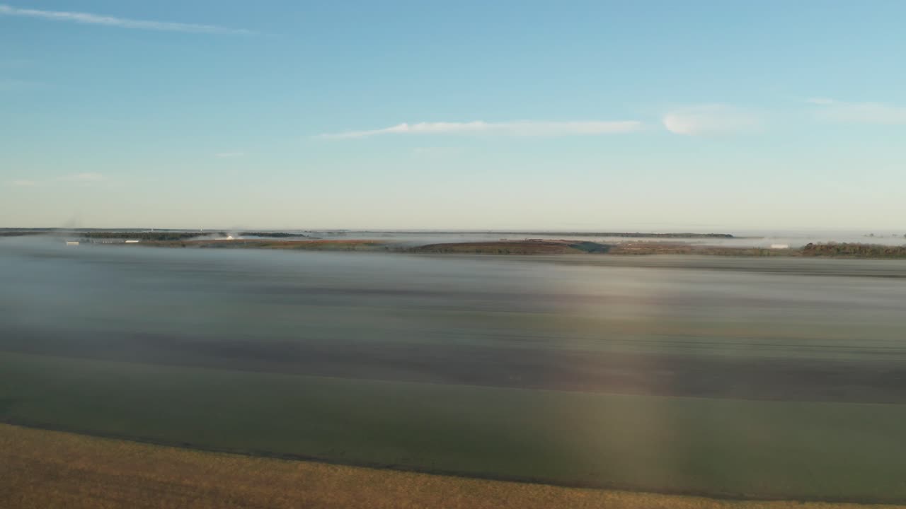 Aerial view of morning light and and fog on farm fields with bright blue sky and soft white clouds overhead. Colorful landscape of farmland and crops. Slide right.