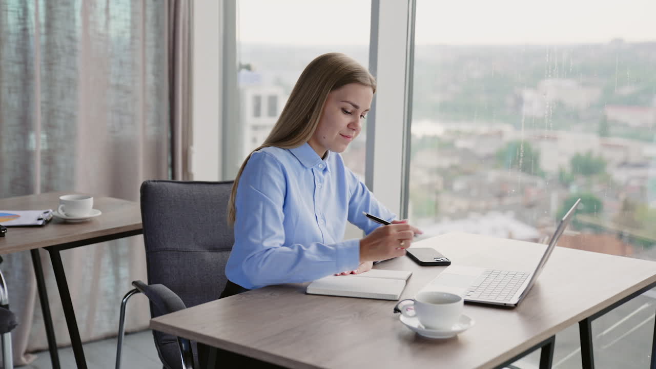 Young businesswoman sitting at desk writing down something into her paper notebook. Panoramic windows with cityscape in blur at backdrop.