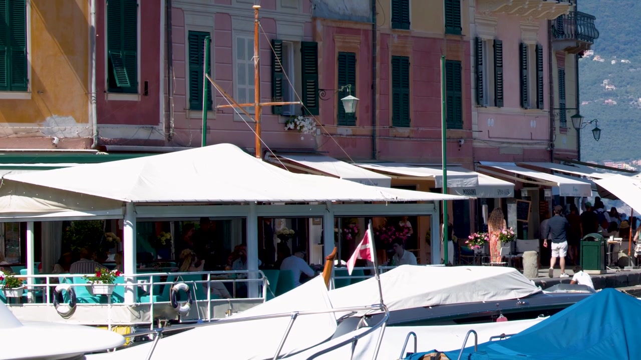 Inquadratura fissa di colorata architettura mediterranea che si affaccia su un porto trafficato con tavoli all'aperto di ristoranti.