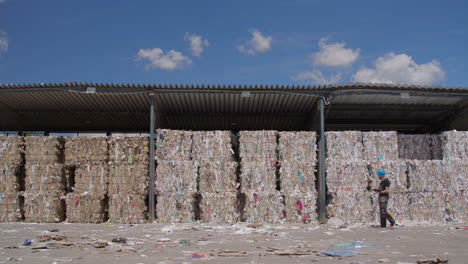 wide static slomo of worker walking by paper bales at recycling plant