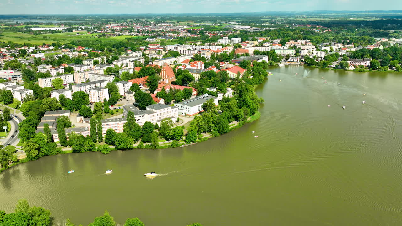 vista aérea de iława, destacando la pintoresca orilla del lago, la exuberante vegetación y las áreas residenciales de la ciudad con edificios históricos, proporcionando un pintoresco paisaje de verano