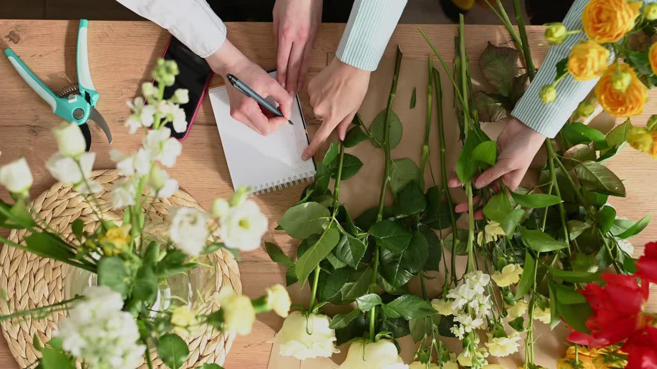 Wanita tak dikenal membuat catatan sambil menerima pesanan dan orang lain merangkai karangan bunga segar di toko bunga.