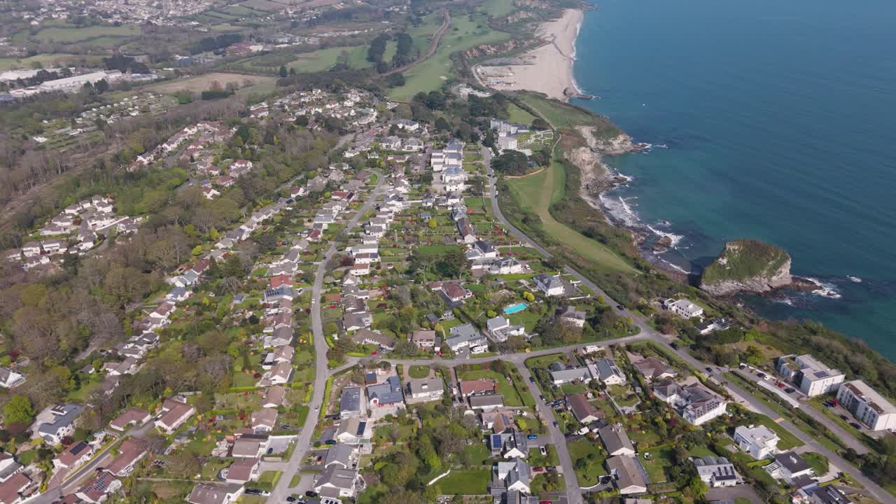Aerial sweep over curved suburban lanes of detached seaside homes with gardens descending toward sandy bay and glimmering sea on the Cornish coast