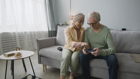 middle aged arabic woman explaining to a senior man how to use the game controller while sitting together on couch at home 2