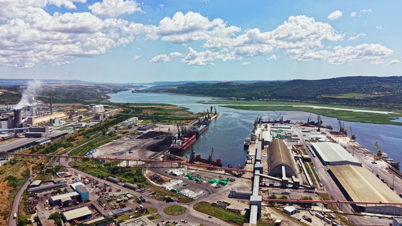 Vista di un trafficato porto industriale con navi ormeggiate e un fiume che scorre vicino a colline verdi sotto un cielo blu con nuvole sullo sfondo