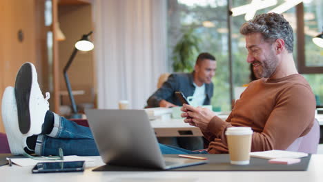 Casually-Dressed-Mature-Businessman-With-Feet-On-Desk-In-Office-Using-Mobile-Phone