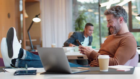 casually dressed mature businessman with feet on desk in office using mobile phone