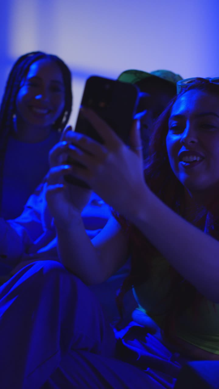 Vertical Video Studio Shot Of Vertical Video Of Group Of Gen Z Friends Sitting On Sofa Posing For Selfie On Mobile Phone At Night With Flashing Light