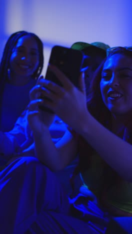 Foto-De-Estudio-De-Vídeo-Vertical-De-Un-Grupo-De-Amigos-De-La-Generación-Z-Sentados-En-Un-Sofá-Posando-Para-Un-Selfie-En-Un-Teléfono-Móvil-Por-La-Noche-Con-Luz-Intermitente