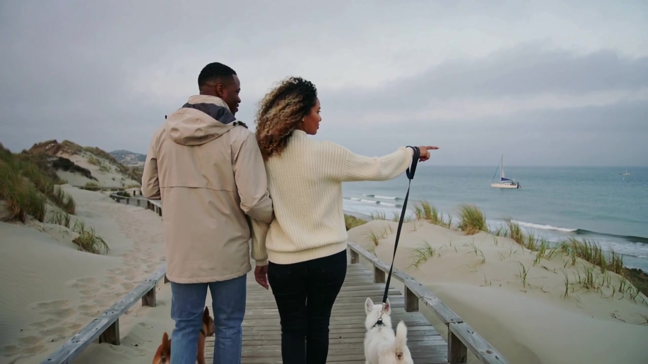 Casal caminhando com jaqueta bege no calçadão, mulher avistando veleiro e apontando, segurando cães na coleira