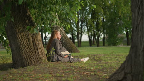 lady in grey clothing sits outdoors under tree reading book with hand on page, leg stretched, head slightly bent as she touch her face background features blurred view of someone walking in distance