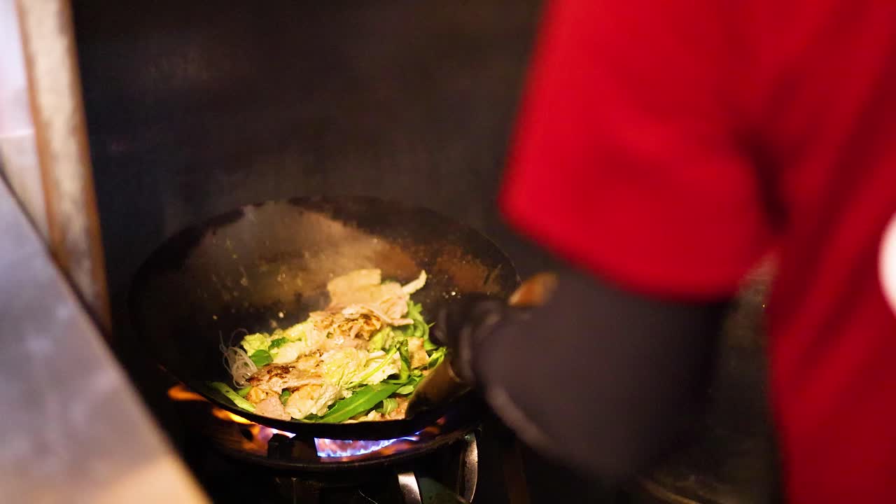 Cook prepares stir-fried sukiyaki in wok over open flame at bustling Bangkok street stall