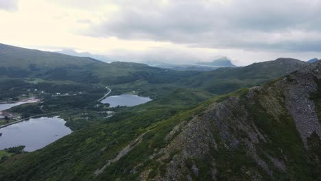 Ein-Hochwinkeliger-Luftblick-über-Einen-Krustigen-Berg-Ein-Dorf-Im-Tal-Mit-Dunklen-Seen-An-Einem-Bewölkten-Tag