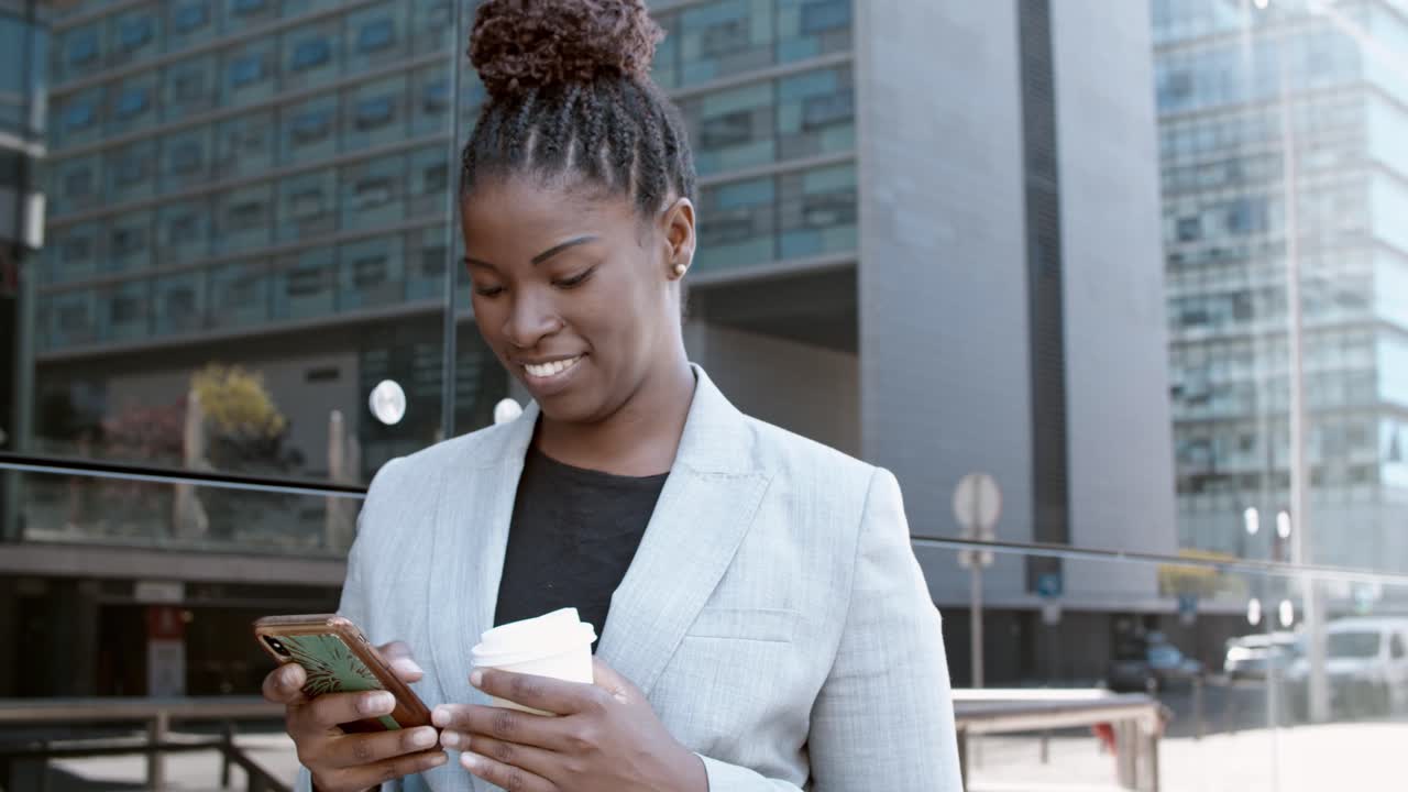 Medium shot of a smiling African-American businesswoman standing outside with paper cup of coffee and texting message on mobile phone
