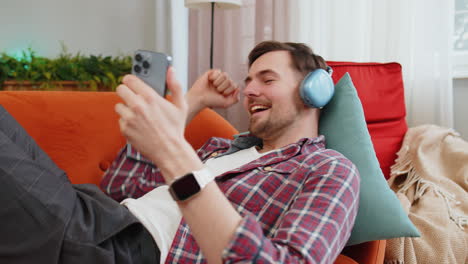 Man-in-wireless-headphones-listening-energetic-dancing-music-on-smartphone-relaxing-lying-on-sofa