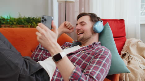 man in wireless headphones listening energetic dancing music on smartphone relaxing lying on sofa