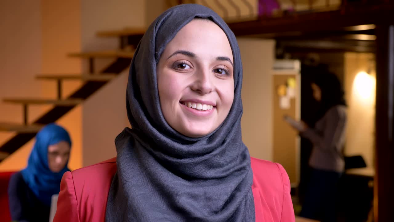 retrato en primer plano de una joven mujer de negocios musulmana positiva con hijab gris mirando directamente a la cámara y sonriendo alegremente en el lugar de trabajo