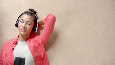 Smiling-biracial-teenage-girl-in-headphones-lying-on-floor-using-smartphone,-copy-space,-slow-motion