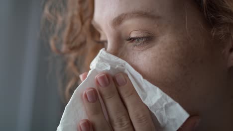 close up of young caucasian  red head woman sneezing and blowing nose into tissue at home