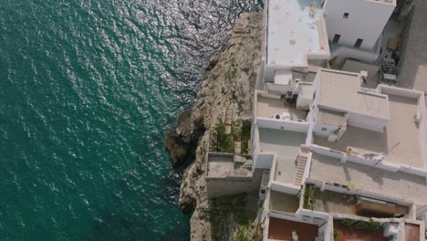 Aerial-pan-up-over-buildings-that-are-sitting-on-cliffs-next-to-the-sea-in-Polignano-a-Mare,-Italy