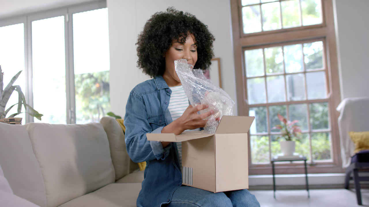 Happy biracial woman unpacking parcel at home, slow motion