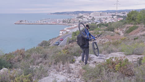 traveller carries his mountain bike on the sholder as group of cyclists pass on a bendy road down below leading to the coastal city