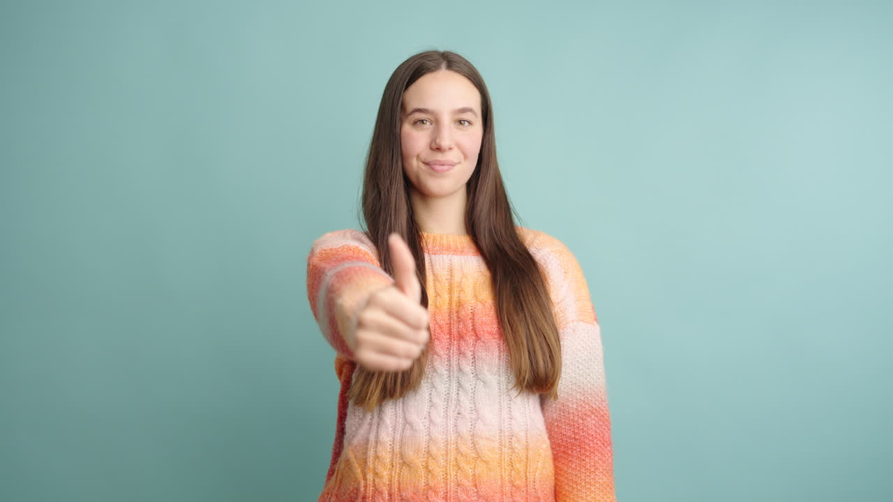 Young woman showing thumbs up and celebrating success on turquoise background