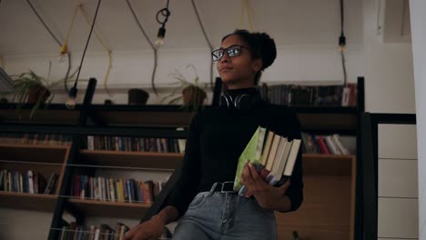 Cheerful-Woman-Standing-Stepping-Down-Stairs-In-Small-Library-Holding-Pile-Of-Books