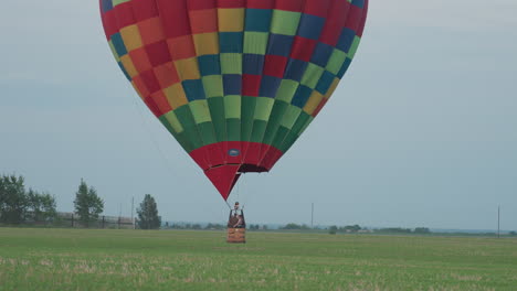 rear view of passenger riding colorful hot air balloon drifting low over expansive green farmland at dusk sky glowing flame illuminating wicker basket and distant trees