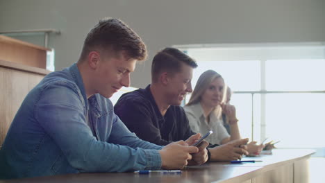 Two-friends-are-sitting-in-a-large-classroom-with-students-and-showing-each-other-funny-videos-from-the-boarding-school.-Have-fun-and-have-fun-at-the-university