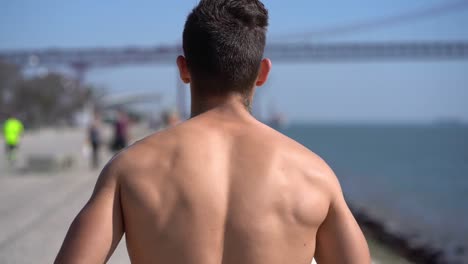 Muscular-shirtless-man-jogging-along-embankment