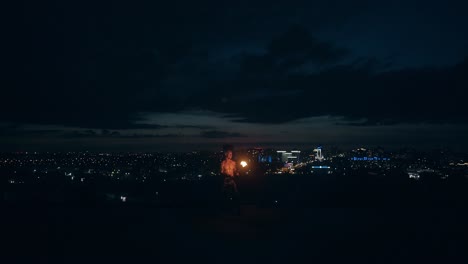 Young-blond-male-breathes-out-fire-makes-fireballs-multiple-times-in-middle-of-night-with-city-skyline-on-black-background