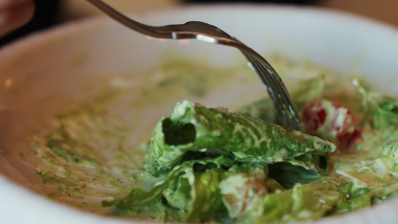 Forchetta che solleva un boccone di lattuga romana cremosa, primo piano di un'insalata condita in una ciotola bianca con pomodorini e crostini. Un condimento lucido ricopre le foglie, una macchia disordinata sul piatto, in un'atmosfera intima da ristorante.