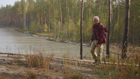 fisherman is walking in forest on high coast of water reservoir carrying rod and backpack resting at nature at autumn vacation