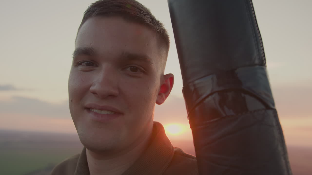 young man stands in hot air balloon basket, warm sunset glow illuminating relaxed face as he turns toward camera, gentle sky fades to soft pastel hues over sprawling farmland horizon
