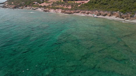 Agua-De-Mar-Turquesa-Con-Olas-En-La-Playa-Y-Turistas-Con-Bote-De-Vacaciones-Durante-El-Verano-En-Cerdeña,-Italia---Toma-Aérea-De-Drones